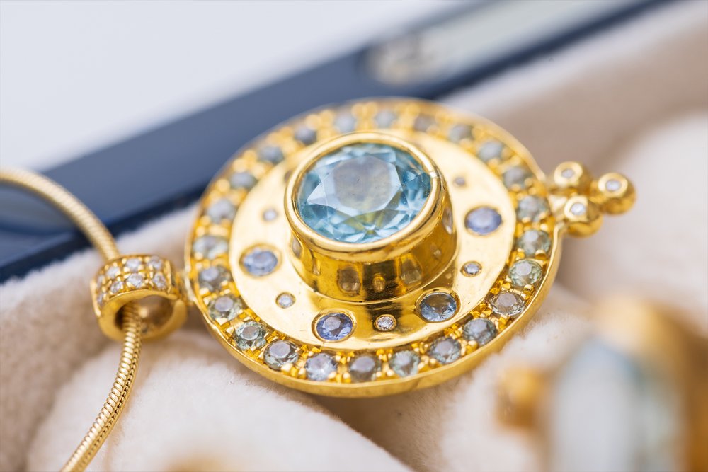 A beautiful mosaic pendant. The Paris & Lily “Dusk” pendant is one-of-a-kind and handcrafted in 22K gold with a 6mm round center aquamarine surrounded by light blue sapphires and diamonds. The pendant has a border of light blue sapphires. Diamonds adorn the chain bail and granules. Shown on a 14K, 16 inch chain.