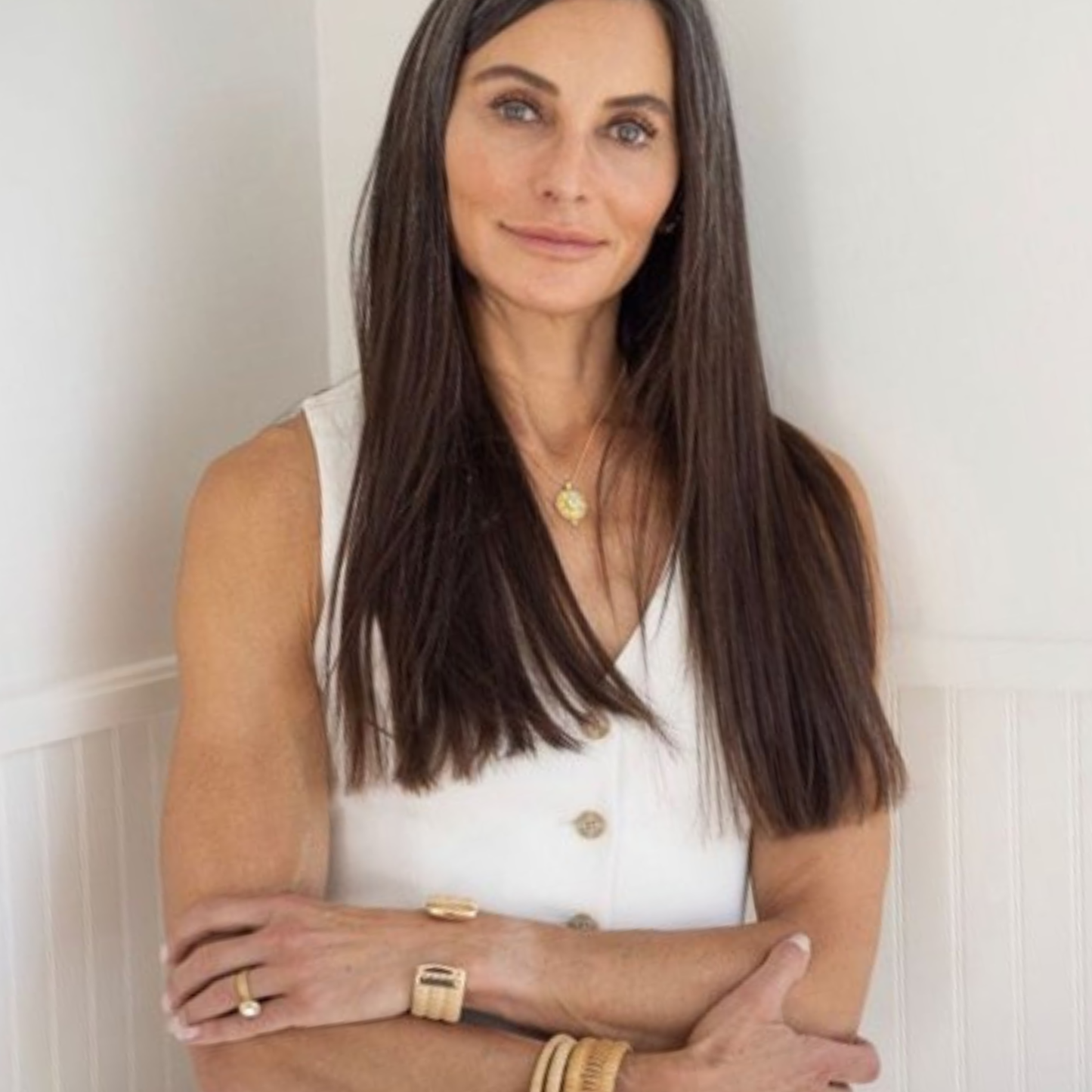 Woman with long hair wearing a white top and gold jewelry and woven cuff bracelets against a light background