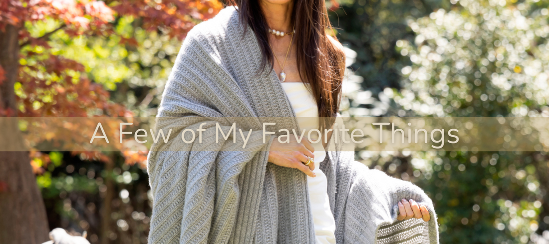 image of brunette female walking barefoot through a beautiful backyard in springtime in jeans and a white tank wrapped in a cashmere ribbed gray blanket. She is wearing pearls.