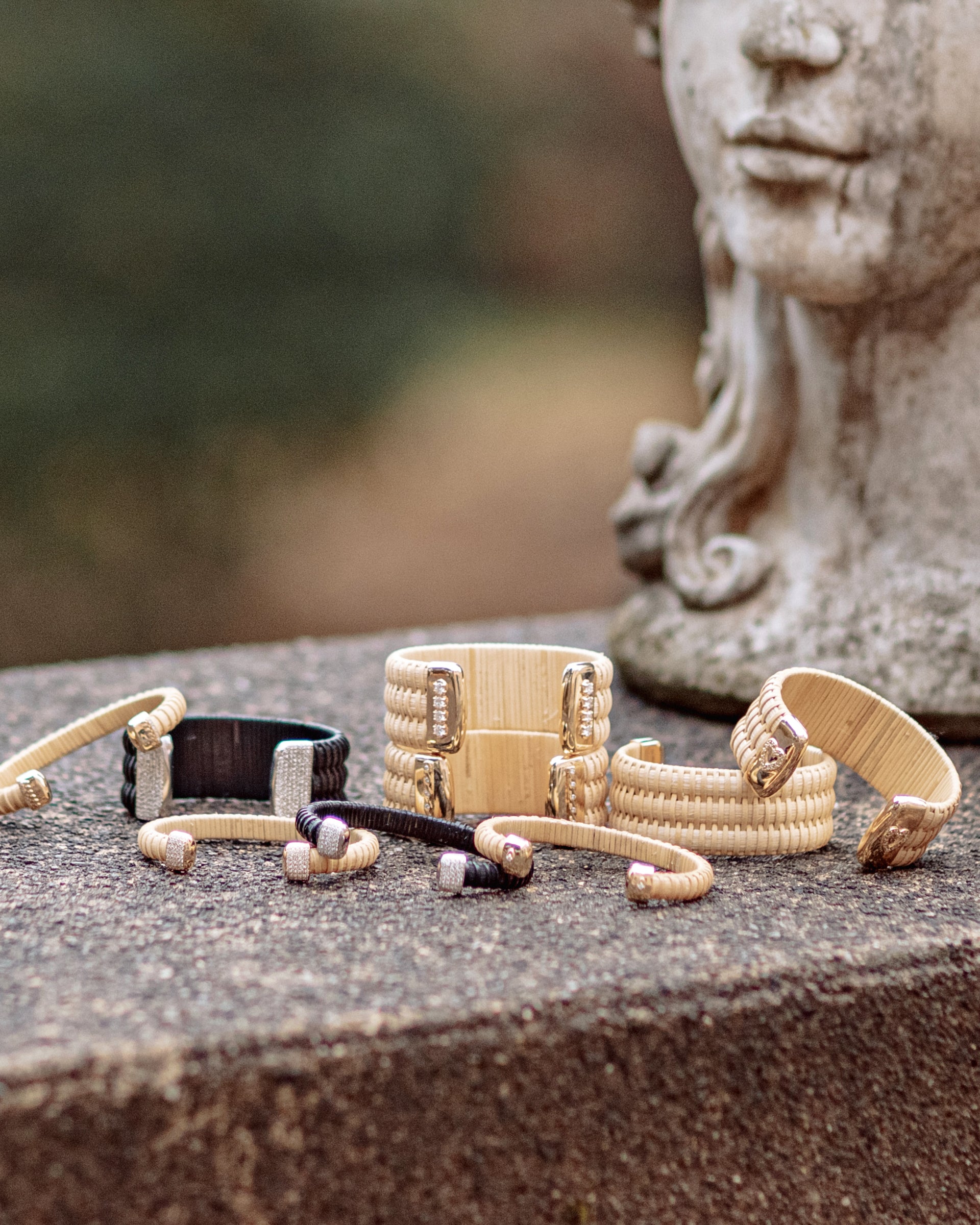 Golden Hour Nantucket Lightship Basket Cuffs, handwoven from natural cane and rattan, with Gold and Diamonds in stack of other cuffs outside on stone next to a statue.
