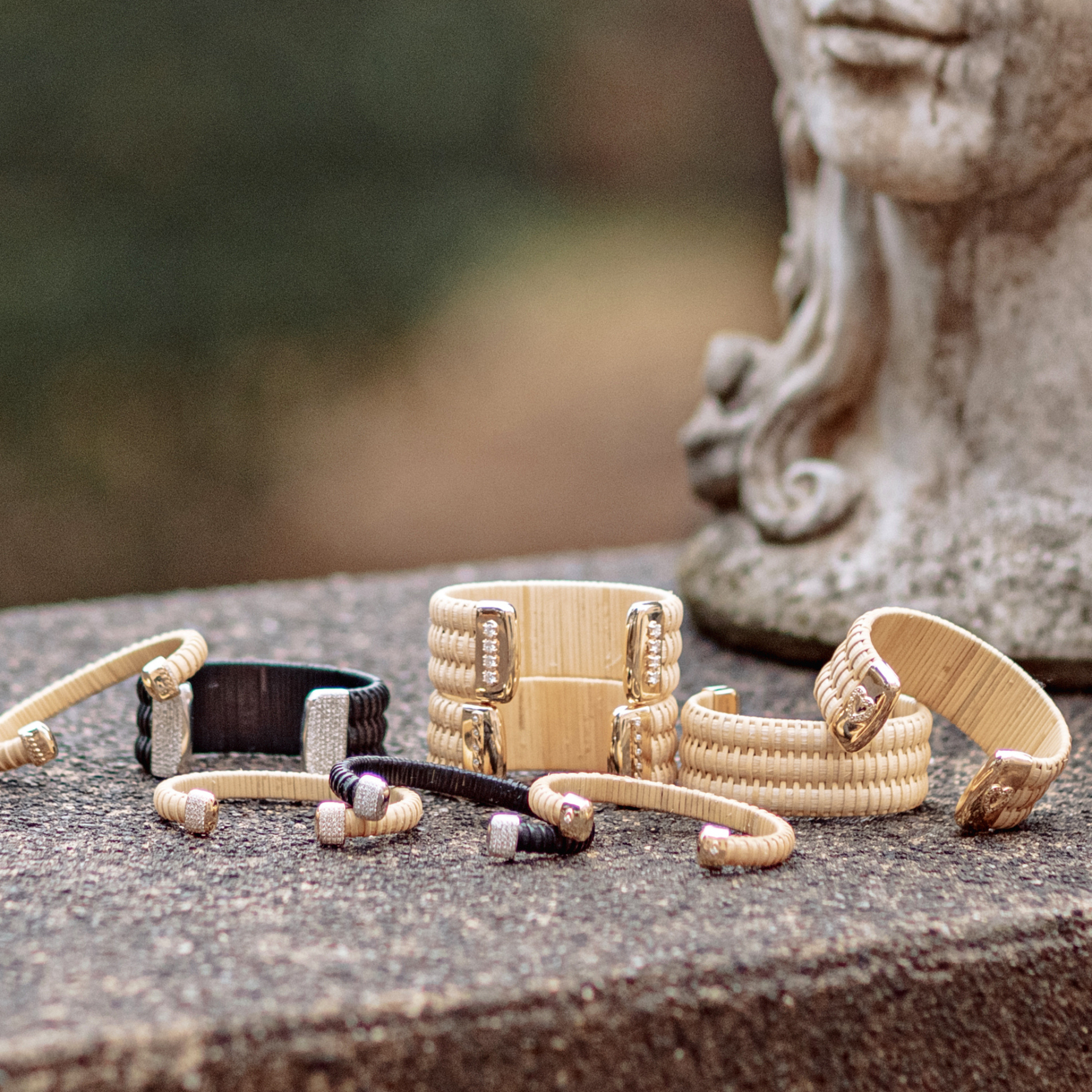 Golden Hour Nantucket Lightship Basket Cuffs, handwoven from natural cane and rattan, with Gold and Diamonds in stack of other cuffs outside on stone next to a statue.