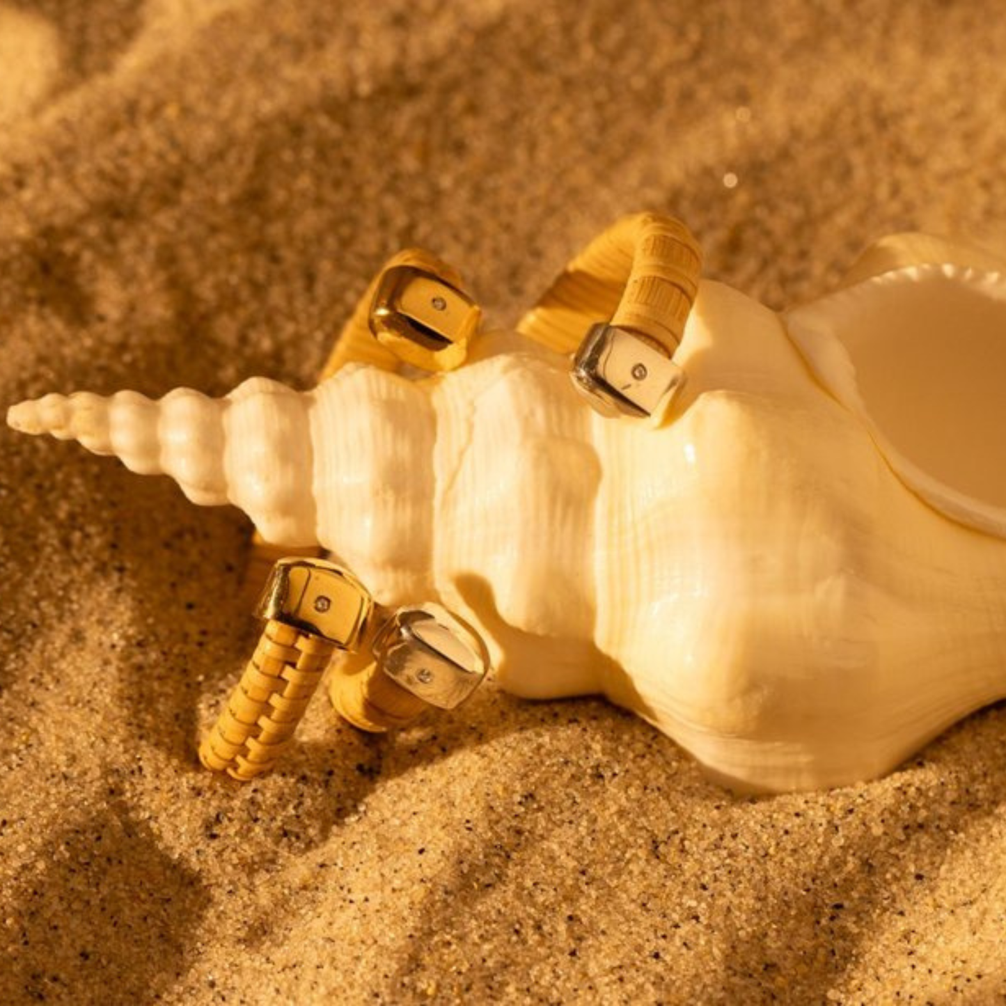 two Nantucket Basket woven cuffs with gold and silver caps atop a unicorn seashell with on a sandy background