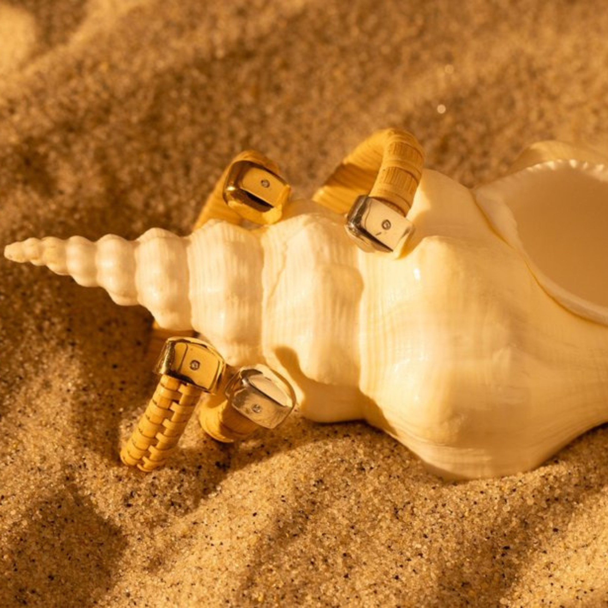 two Nantucket Basket woven cuffs with gold and silver caps atop a unicorn seashell with on a sandy background